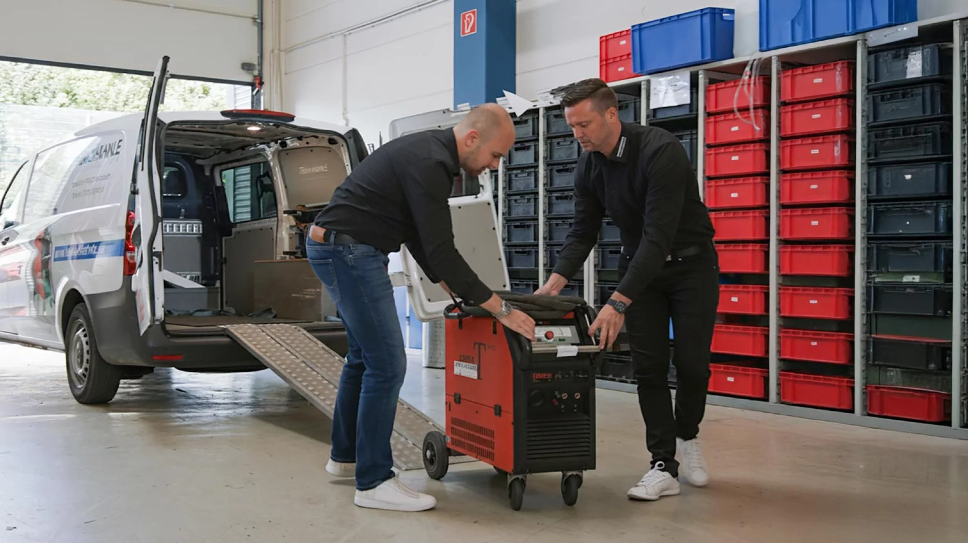 Zwei Mitarbeiter eines SCHWEISS-PROFIS laden eine Schweißstromquelle aus dem Servicefahrzeug vor Regalen mit Kleinteilen
